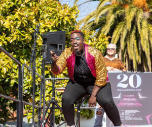 A Black man wearing a sparkly jacket and colorful eyeshadow singing on an outdoor stage