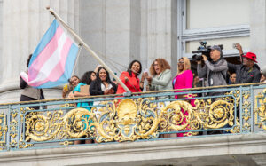 In celebration of Transgender History Month, San Francisco Mayor London Breed and trans activist Donna Personna raise the transgender pride flag outside San Francisco City Hall, surrounded by guests and photographers on the mayor's balcony.